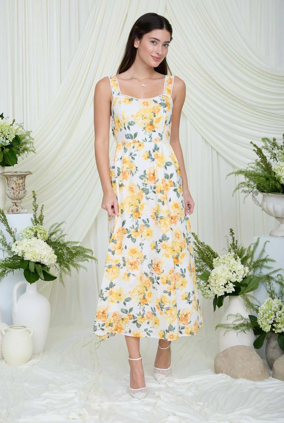 Woman wearing a yellow floral dress standing in a decorated room with white drapes and floral arrangements.