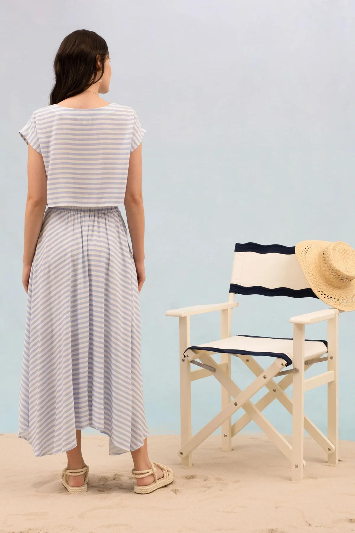 Woman in a striped dress standing next to a director's chair with a straw hat on a light blue background