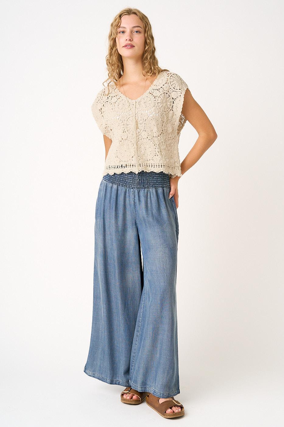 Woman wearing a beige lace top and blue wide-leg pants on a white background