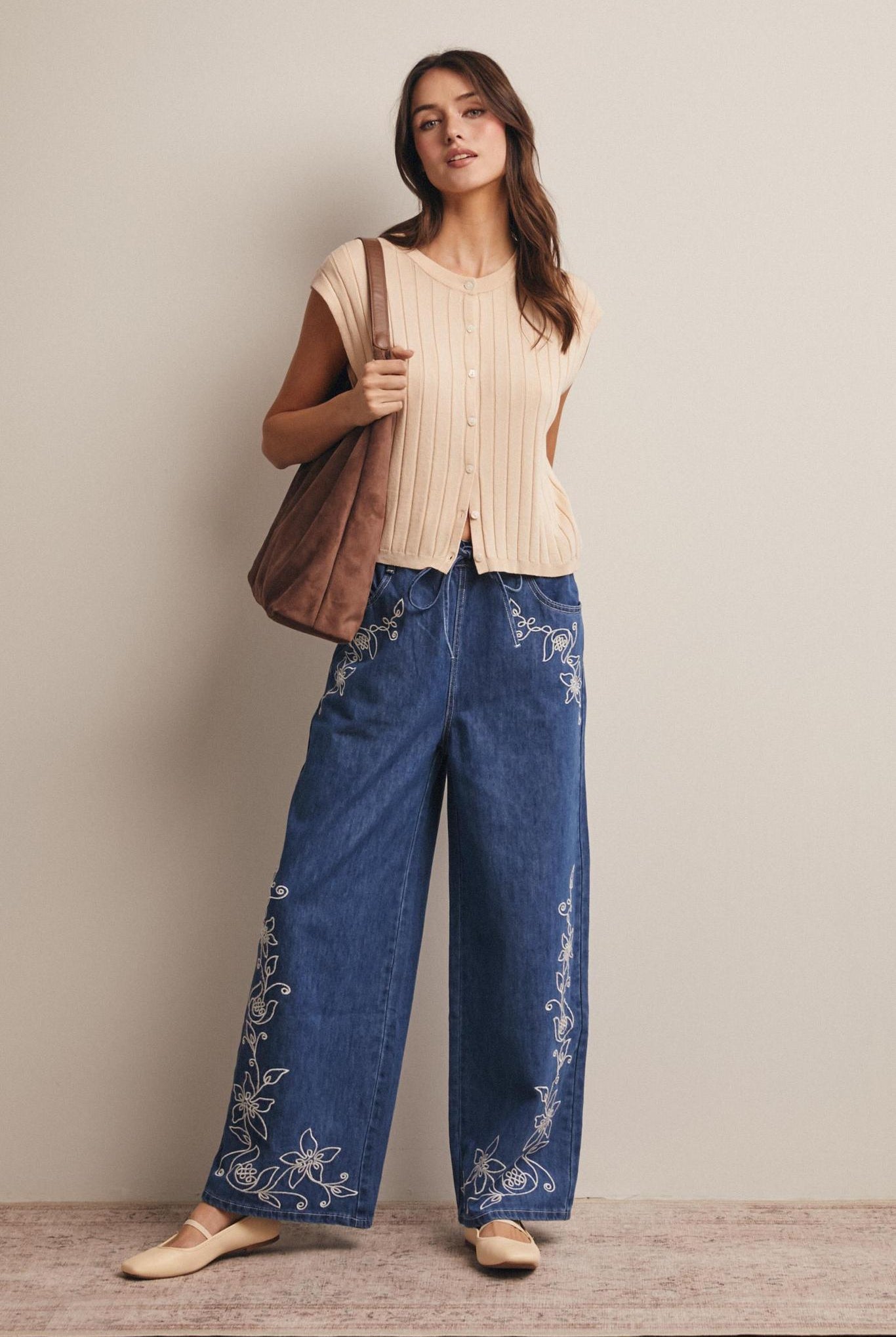 Woman wearing a beige top and blue pants with floral embroidery, holding a brown bag.