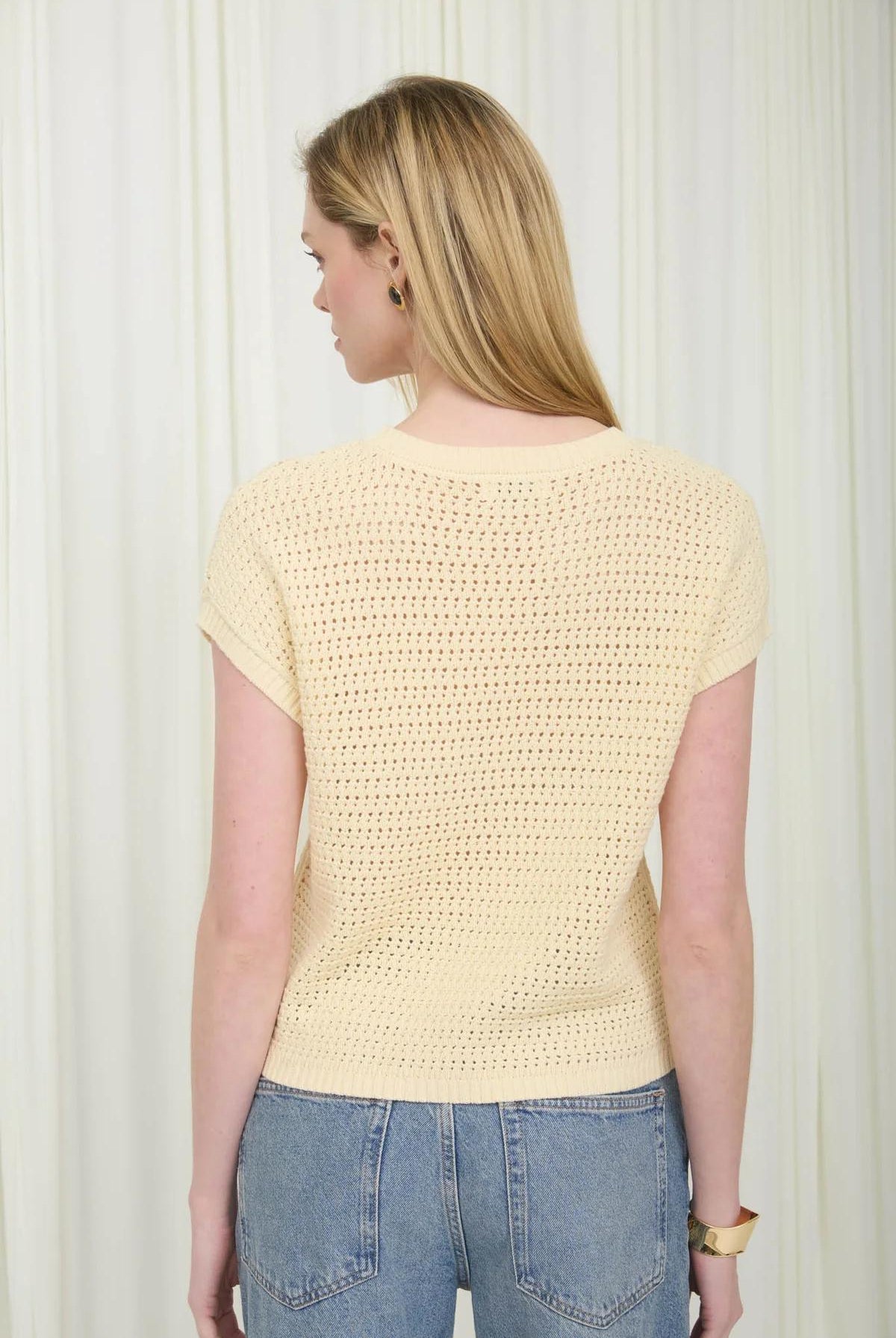 Woman wearing a light yellow crochet top and blue jeans against a white background