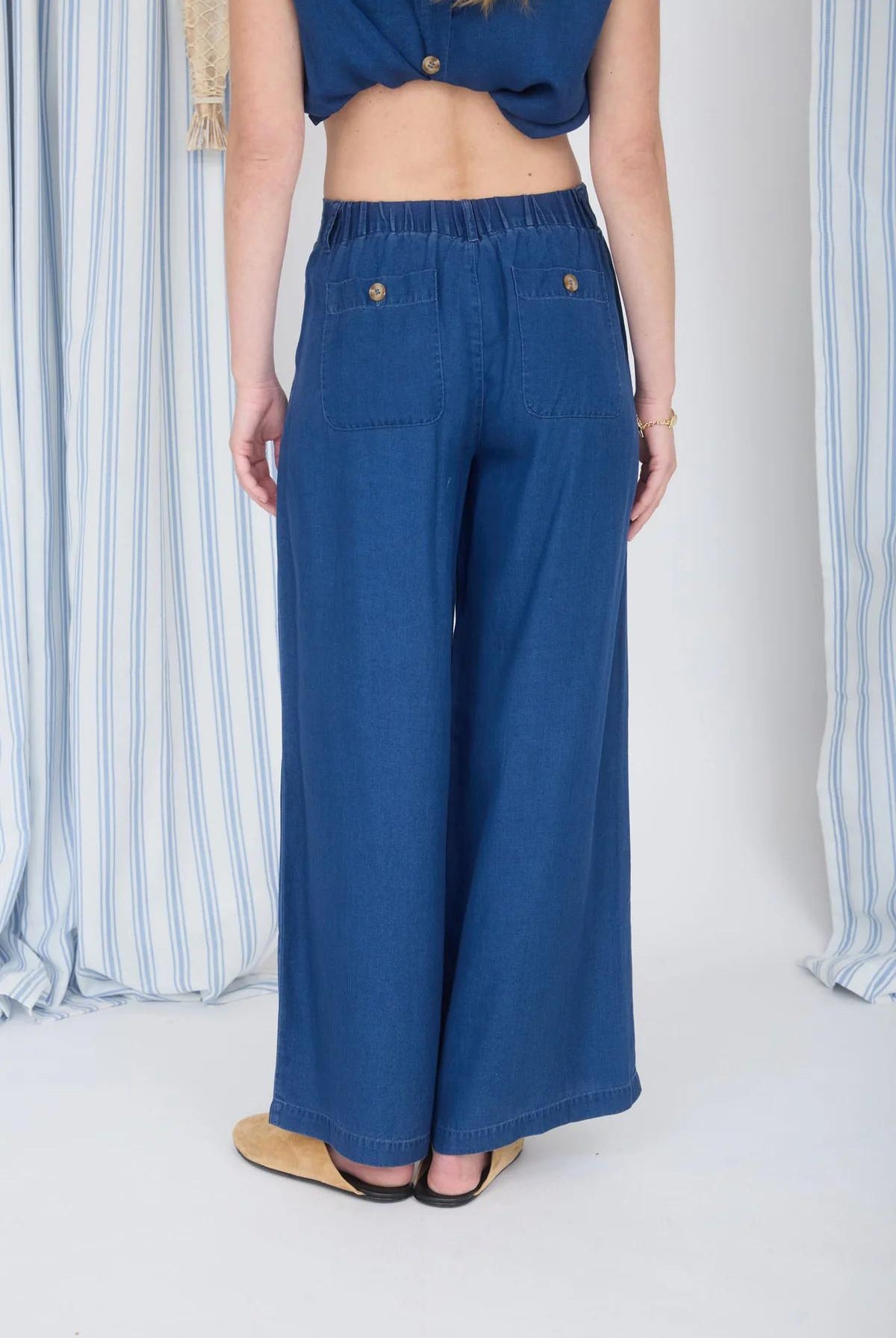 Person wearing blue wide-leg jeans standing against a white background with striped curtains.