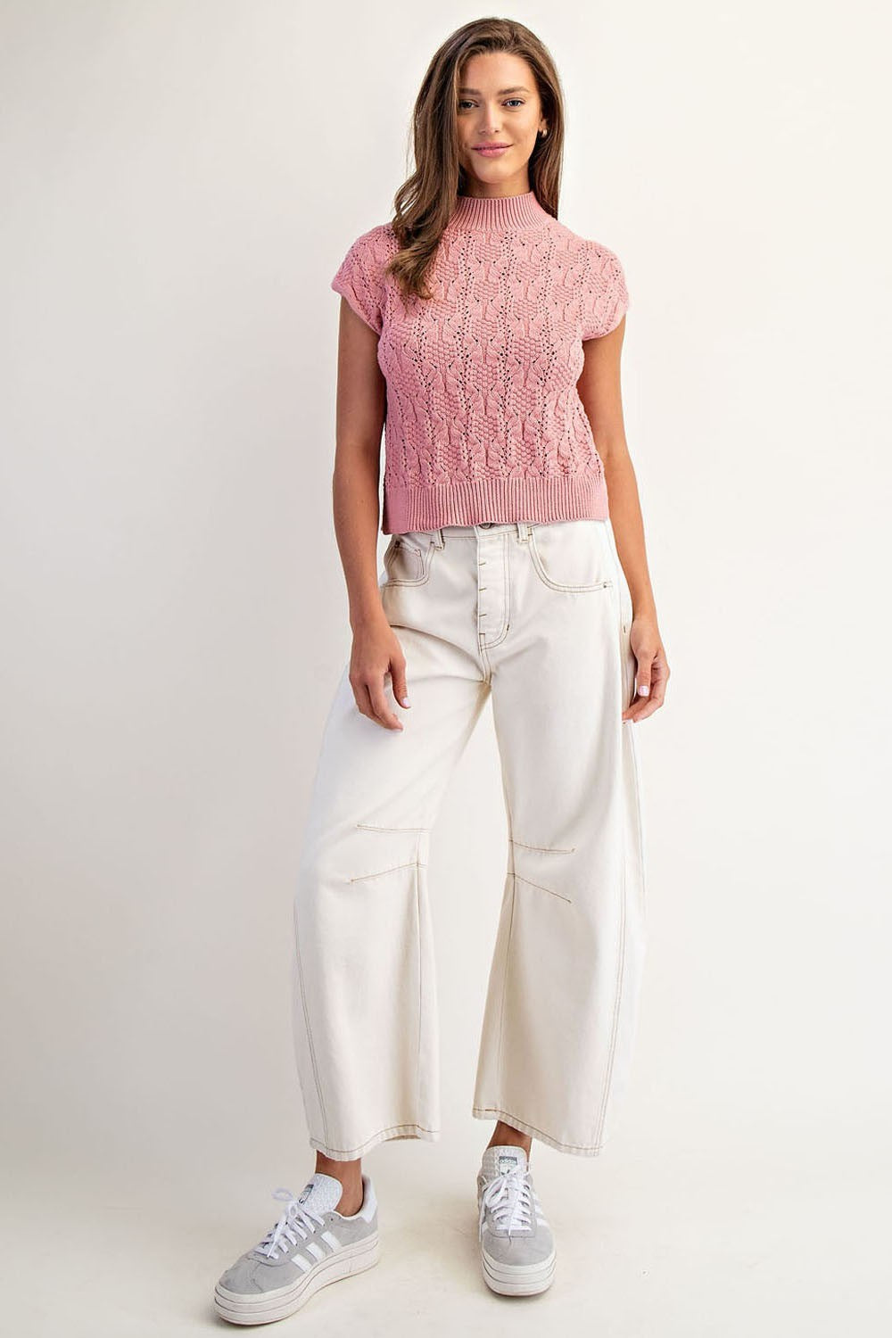 Woman wearing a pink knitted top and white pants on a plain background