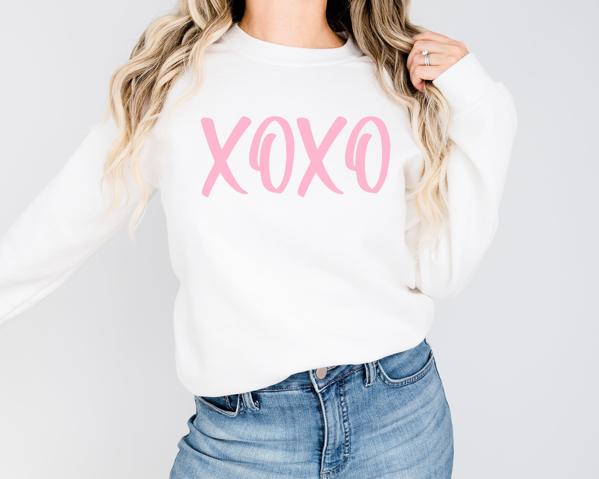 Person wearing a white sweatshirt with pink 'XOXO' text and blue jeans on a light gray background