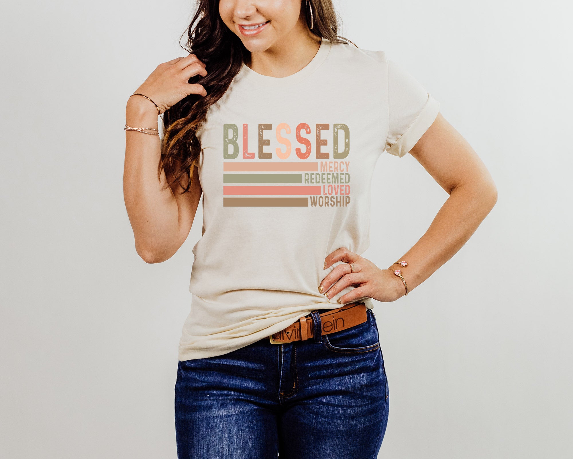 A woman wearing a beige graphic tee with the text 'Blessed, Mercy, Redeemed, Loved, Worship' printed on the front.
