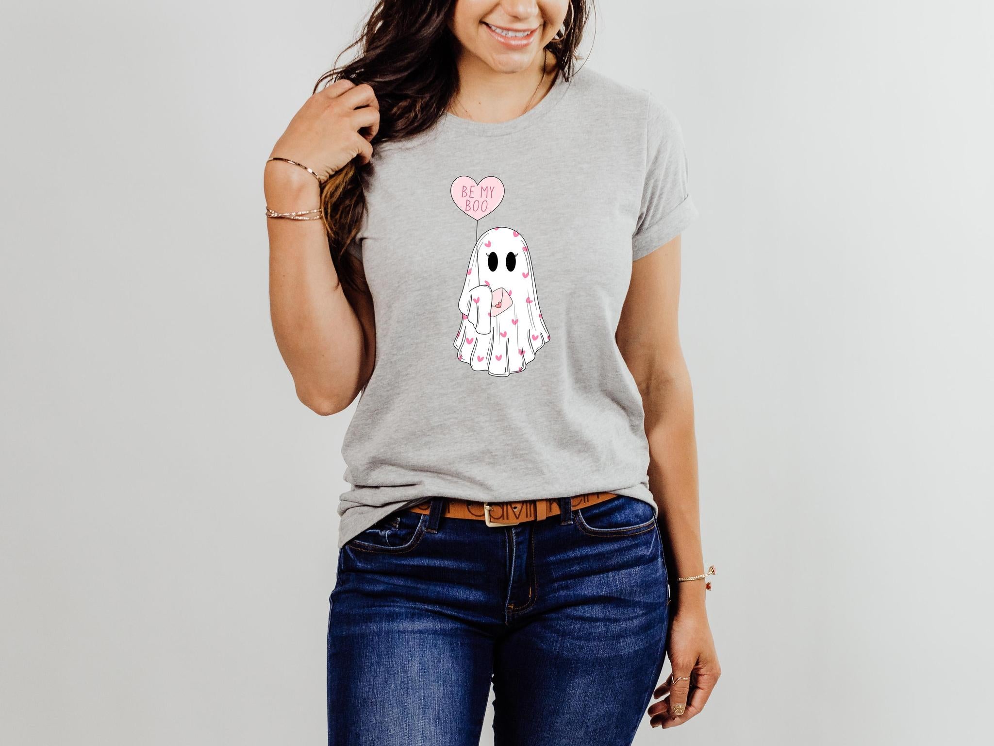 Person wearing a gray t-shirt with a ghost design holding a heart-shaped balloon on a plain background