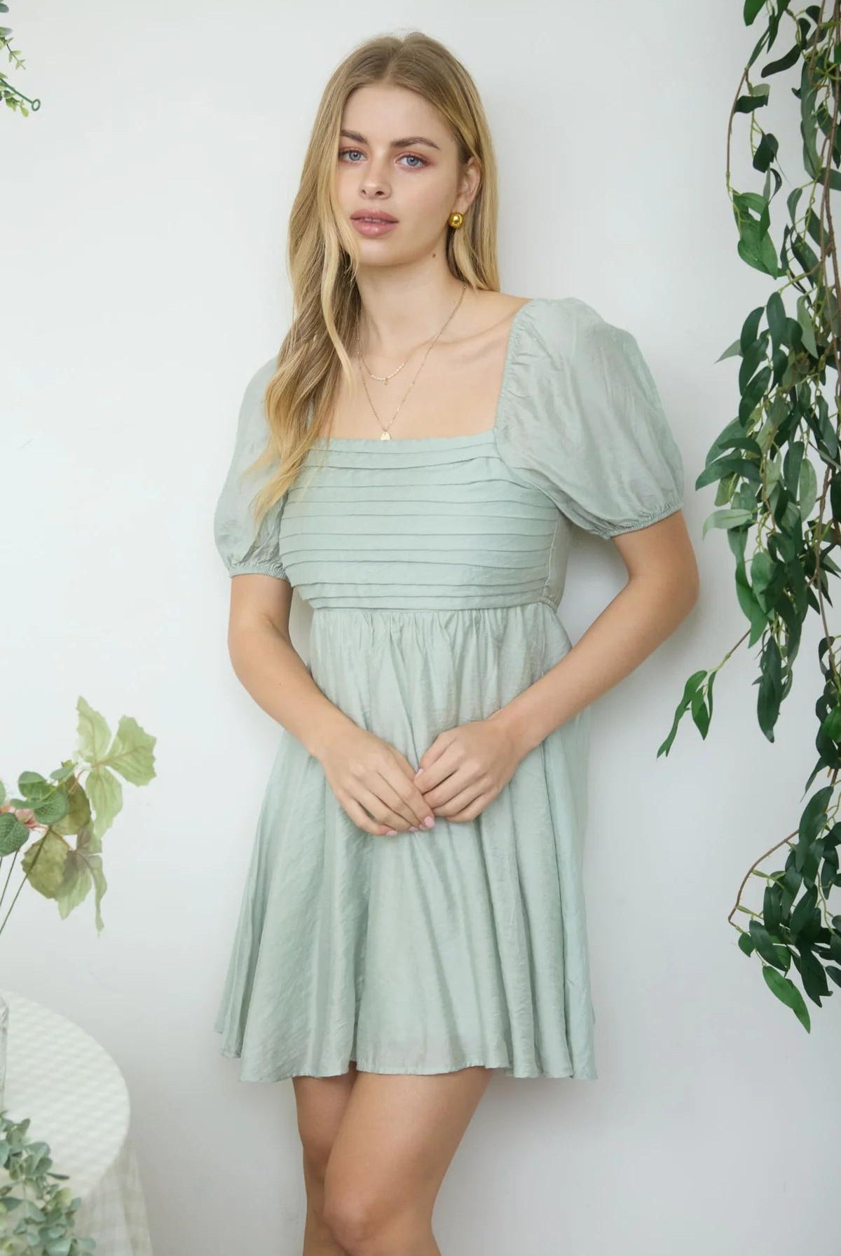 Woman wearing a light green dress standing against a white wall with plants around.
