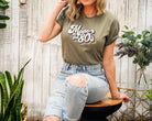 Woman wearing a 'Made in the 80s' t-shirt sitting on a stool with plants around.
