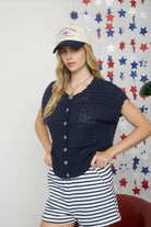 Woman wearing a navy blue cardigan and striped shorts with a cap against a decorative wall.