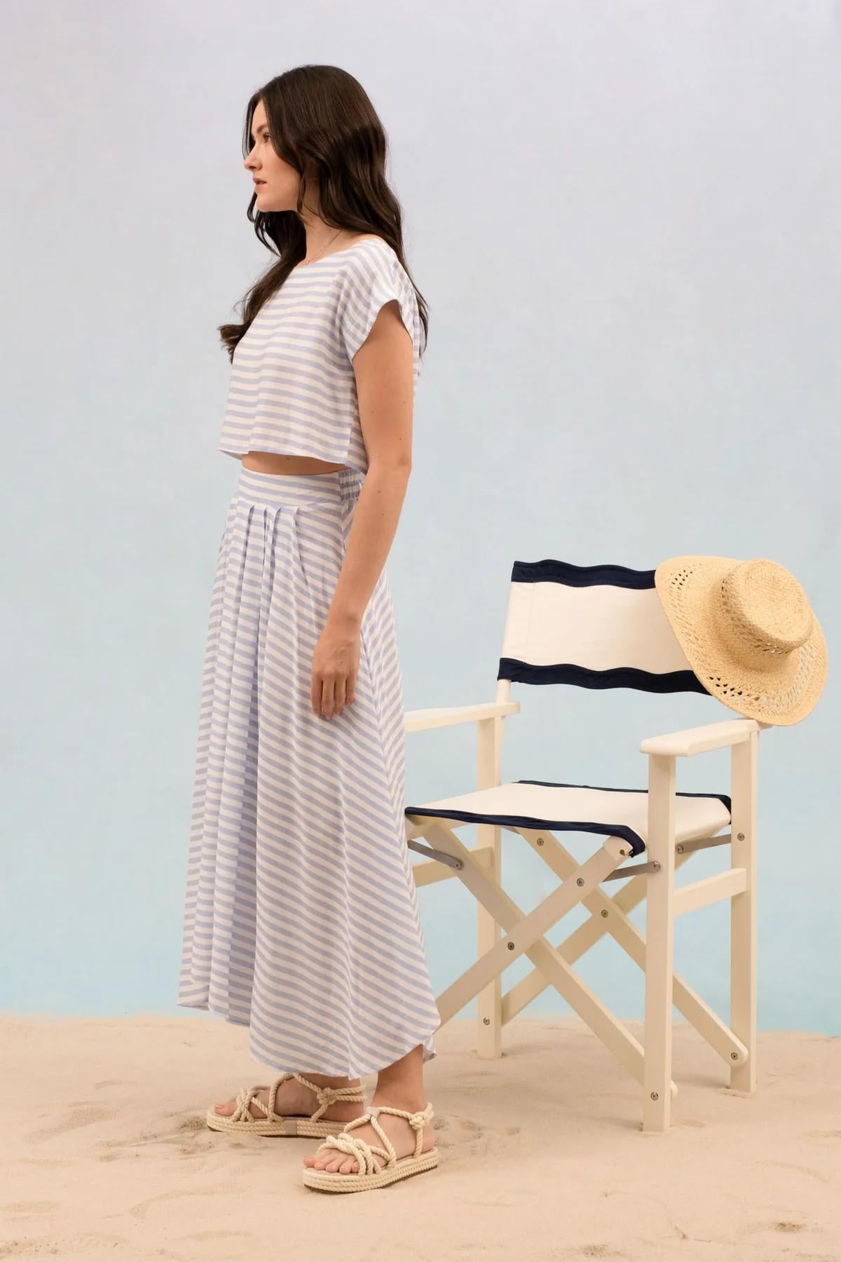Woman in striped outfit standing on a beach with a chair and straw hat in the background.