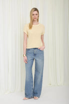 Woman wearing a yellow top and blue jeans standing against a white curtain.