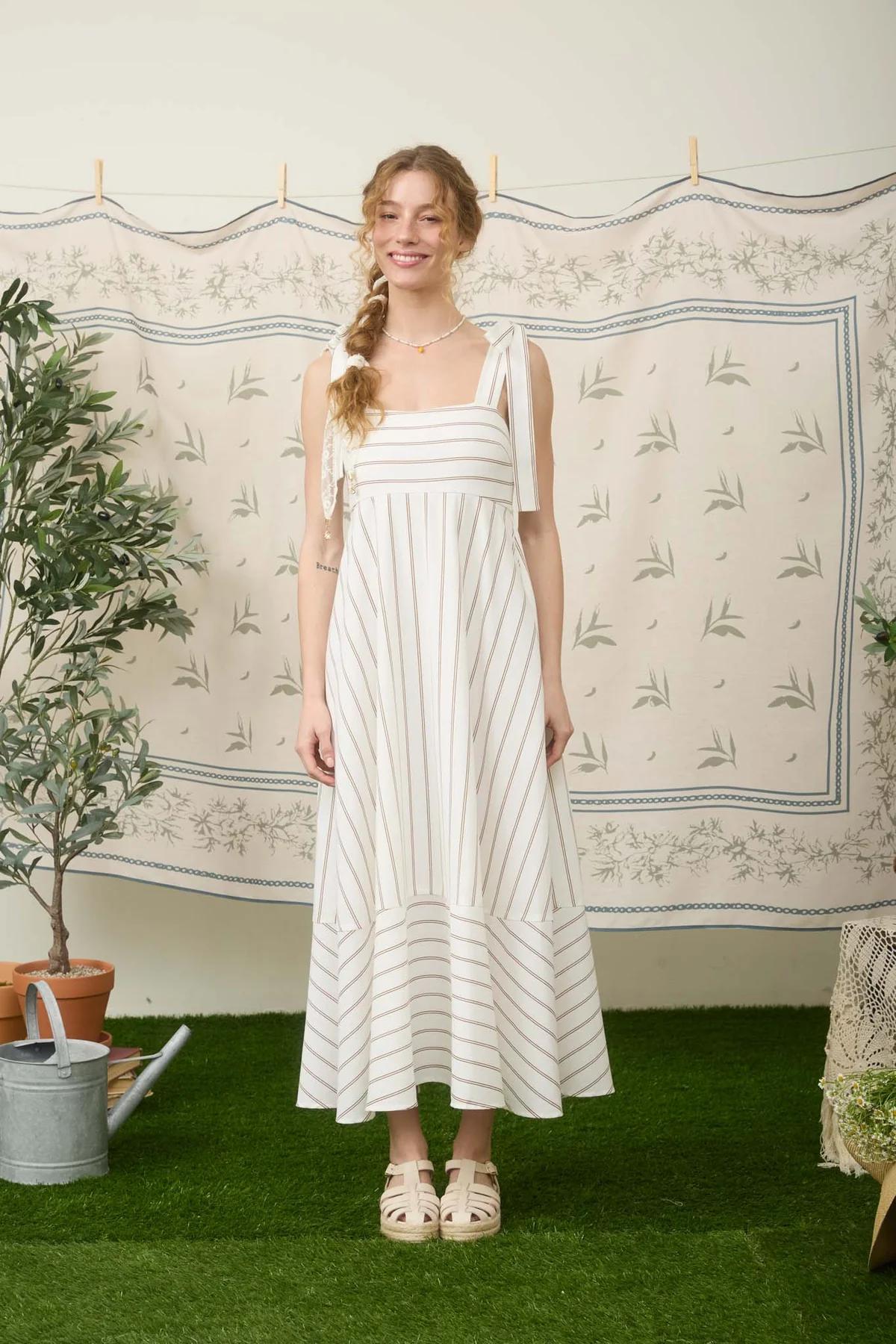 Woman in a striped dress standing in front of a decorative wall with plant and pot on the left.