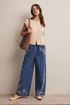 Woman wearing a beige top and blue pants with floral embroidery, holding a brown bag.