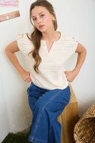 Woman wearing a white top and blue jeans sitting on a wooden stool against a white wall.