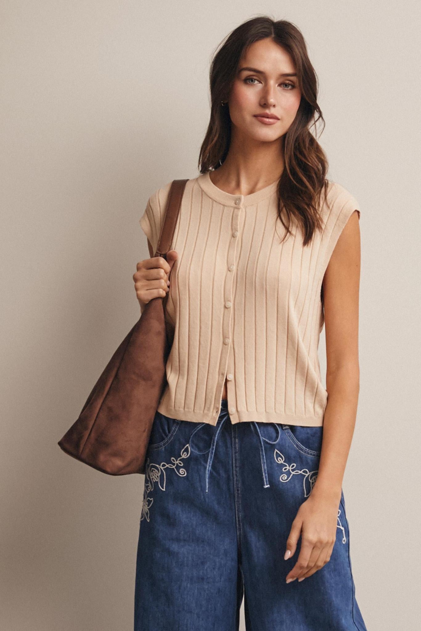 Woman wearing a beige sleeveless top and blue jeans, holding a brown leather bag against a plain background