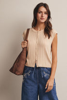 Woman wearing a beige sleeveless top and blue jeans, holding a brown leather bag against a plain background