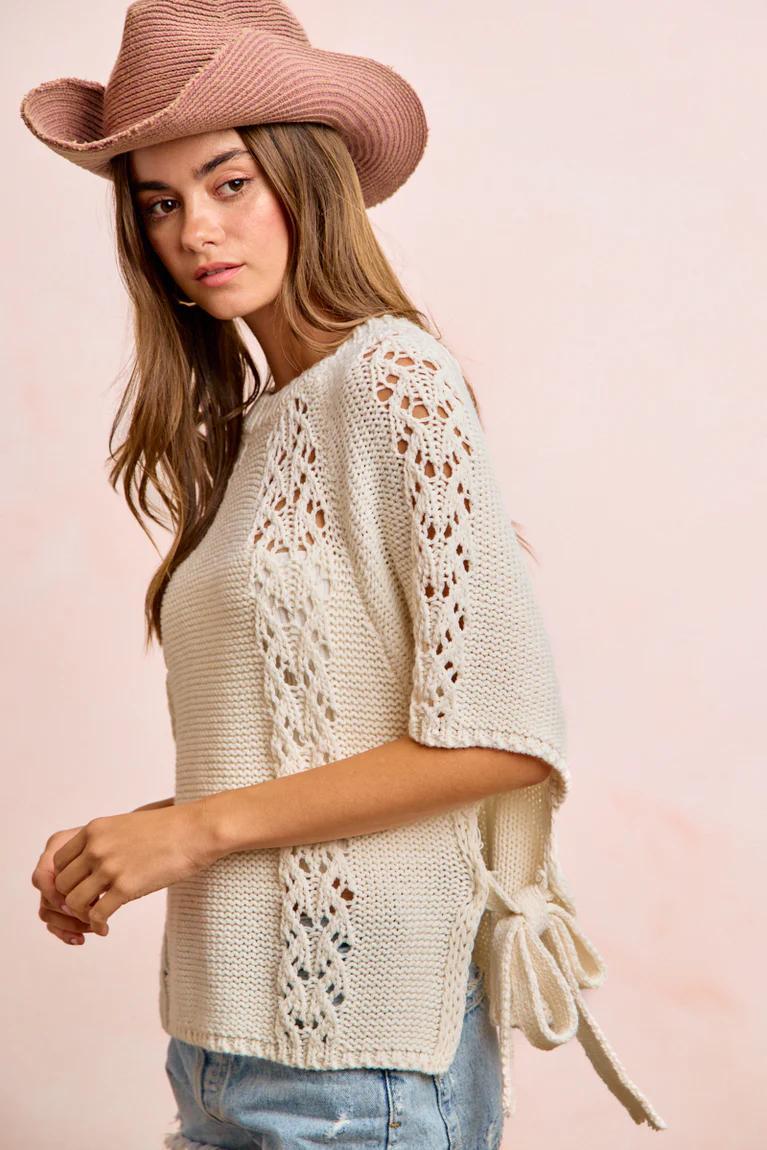 Woman wearing a beige knitted top with fringe details and a pink hat on a light pink background