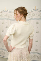 Woman wearing a cream-colored blouse with lace details in a room with floral wallpaper.