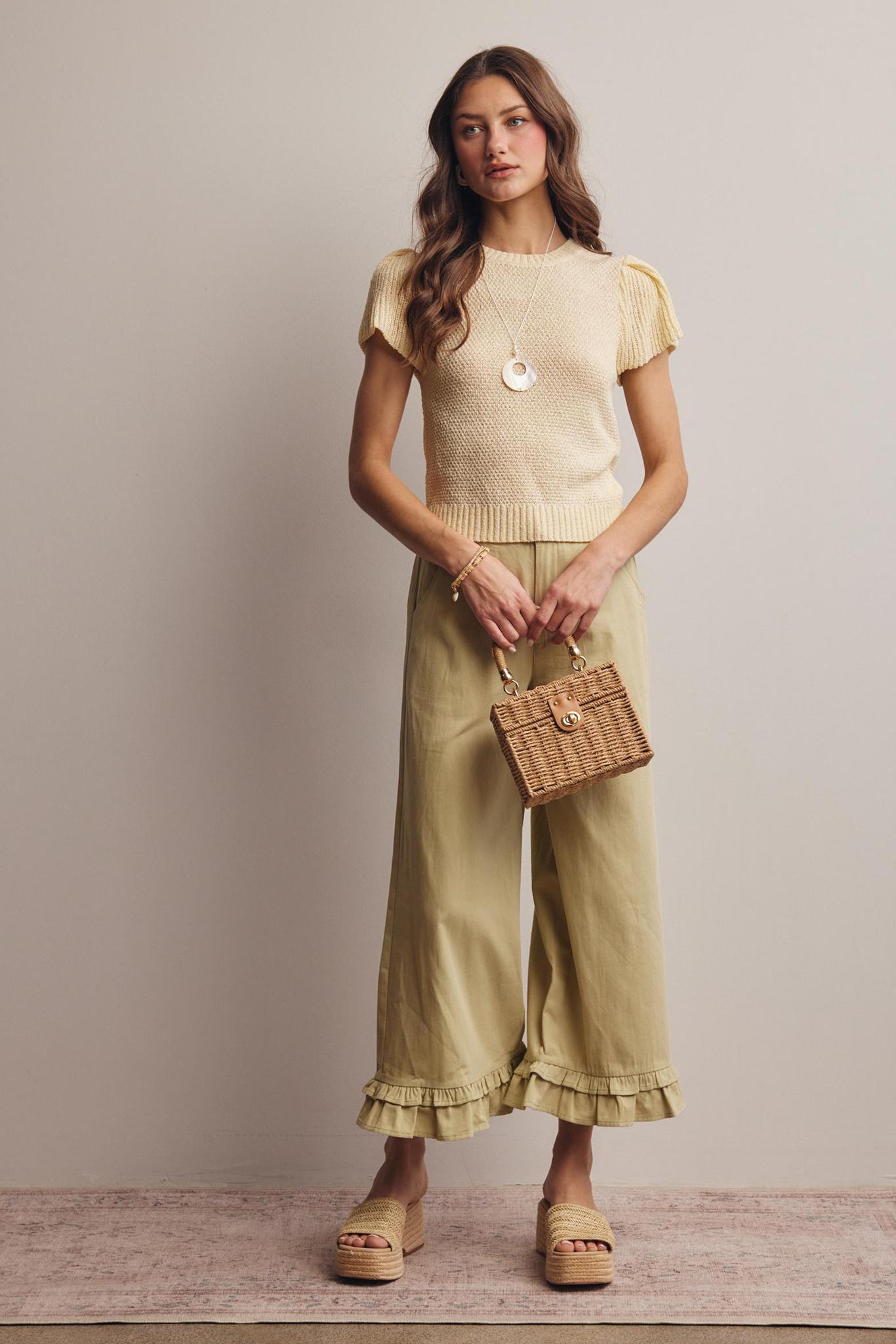Woman wearing a yellow top and green pants holding a woven bag against a neutral background