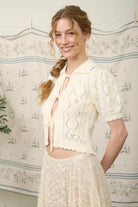 Woman wearing a cream-colored lace outfit in front of a floral curtain.