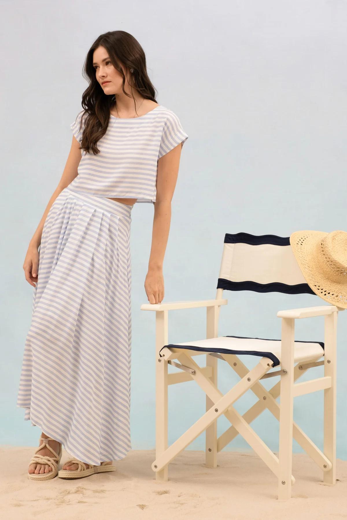 Woman in striped outfit standing next to a director's chair with a straw hat on a light blue background