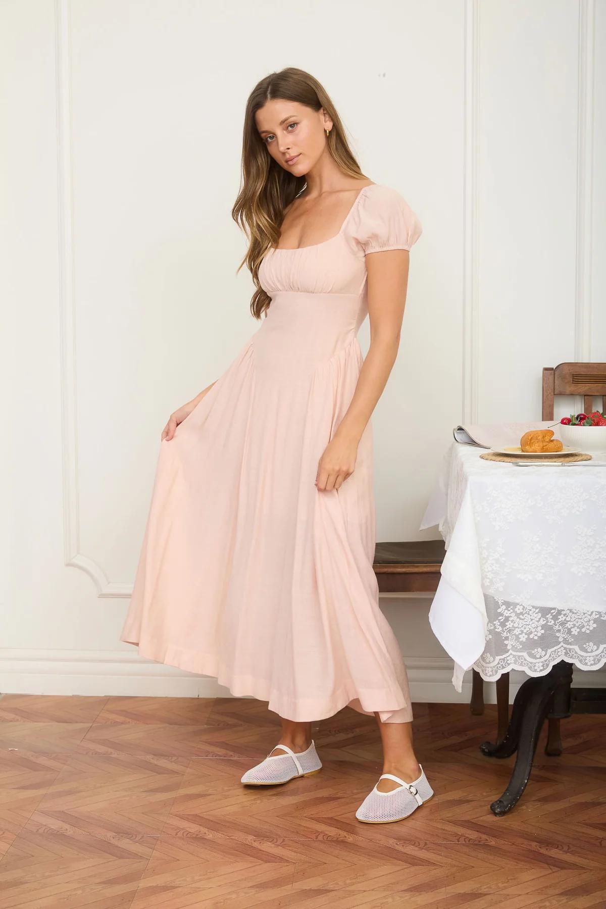 Woman in a pink dress standing in a room with wooden floor and white walls.