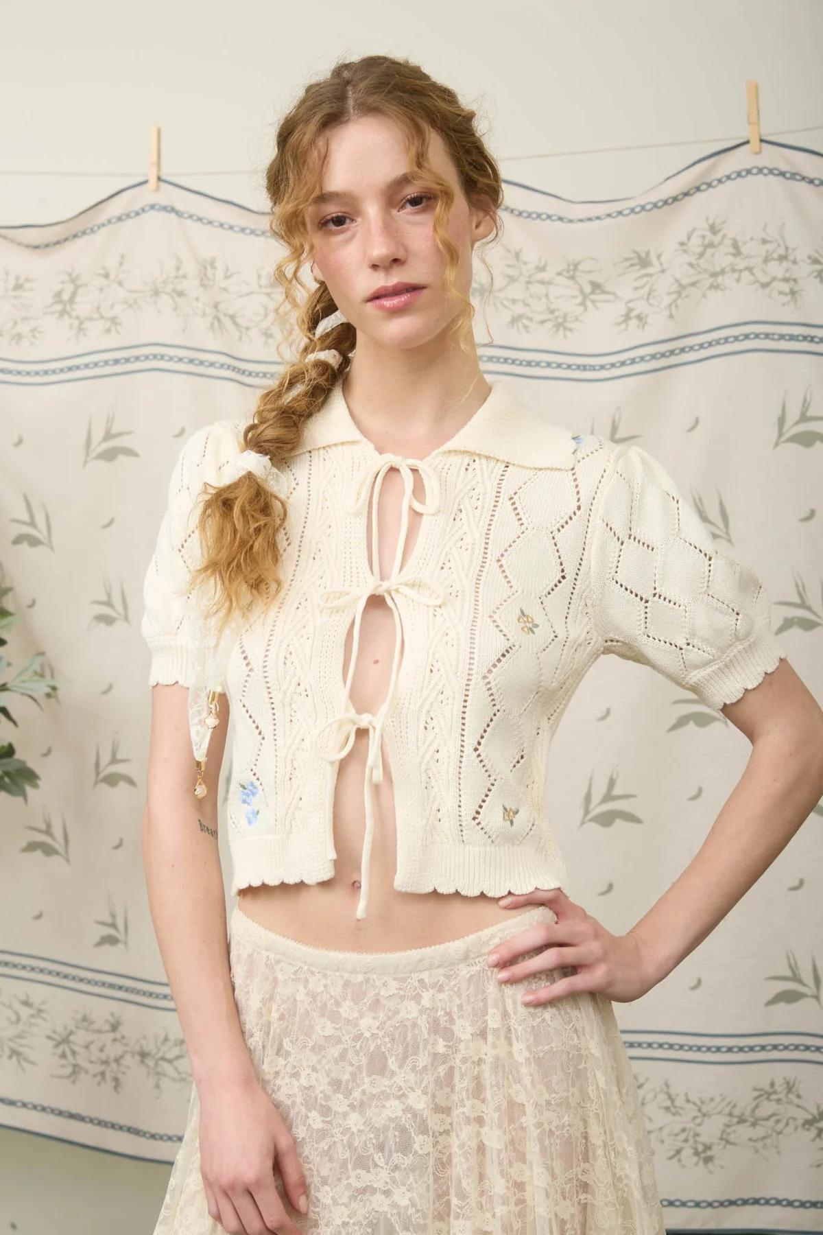 Woman wearing a cream-colored lace top and skirt against a floral-patterned wall.