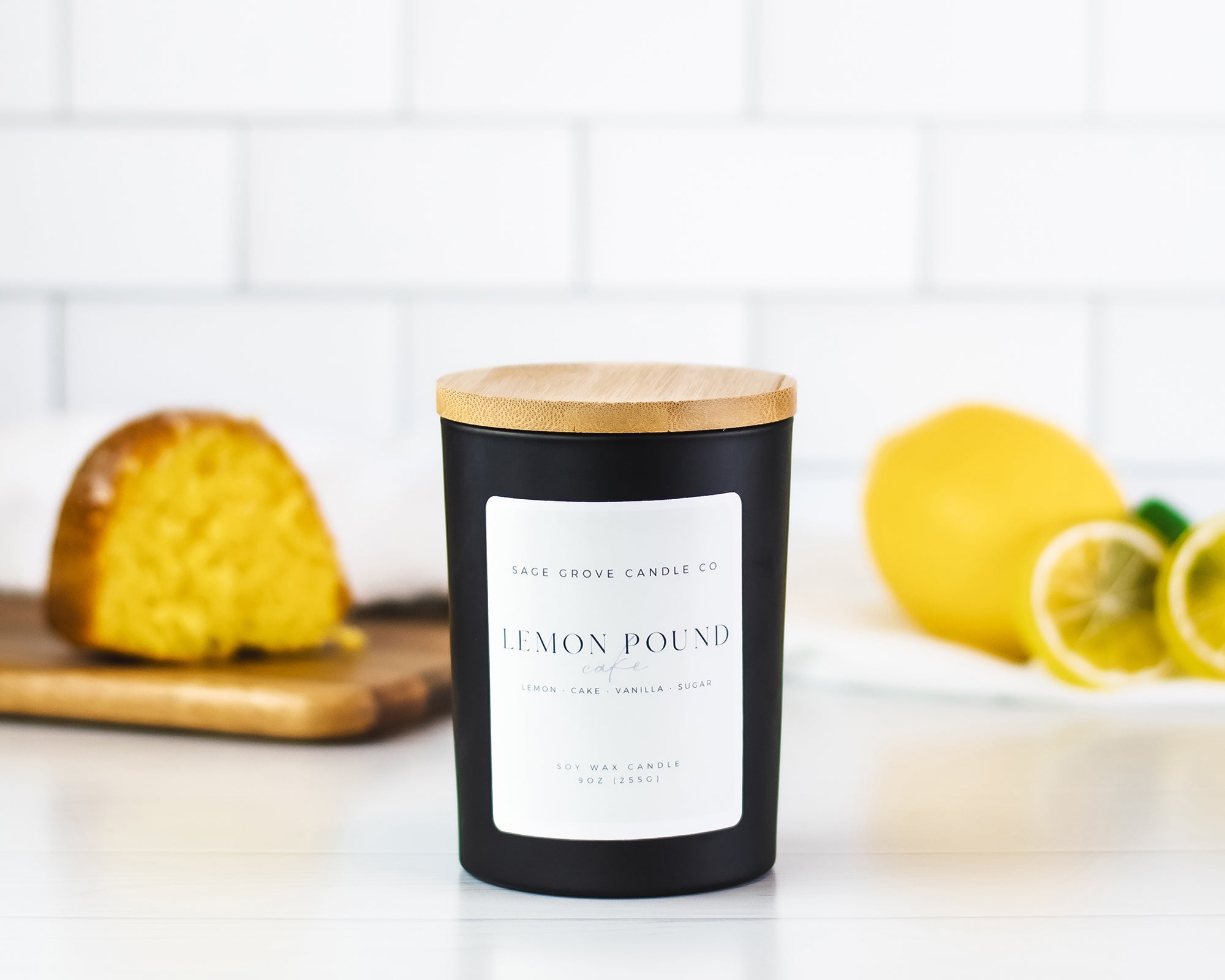 Black candle with a wooden lid labeled 'Lemon Pound' on a white surface with lemons and a cake in the background.