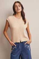 Woman wearing a beige sleeveless ribbed top and blue jeans with floral embroidery on a plain background