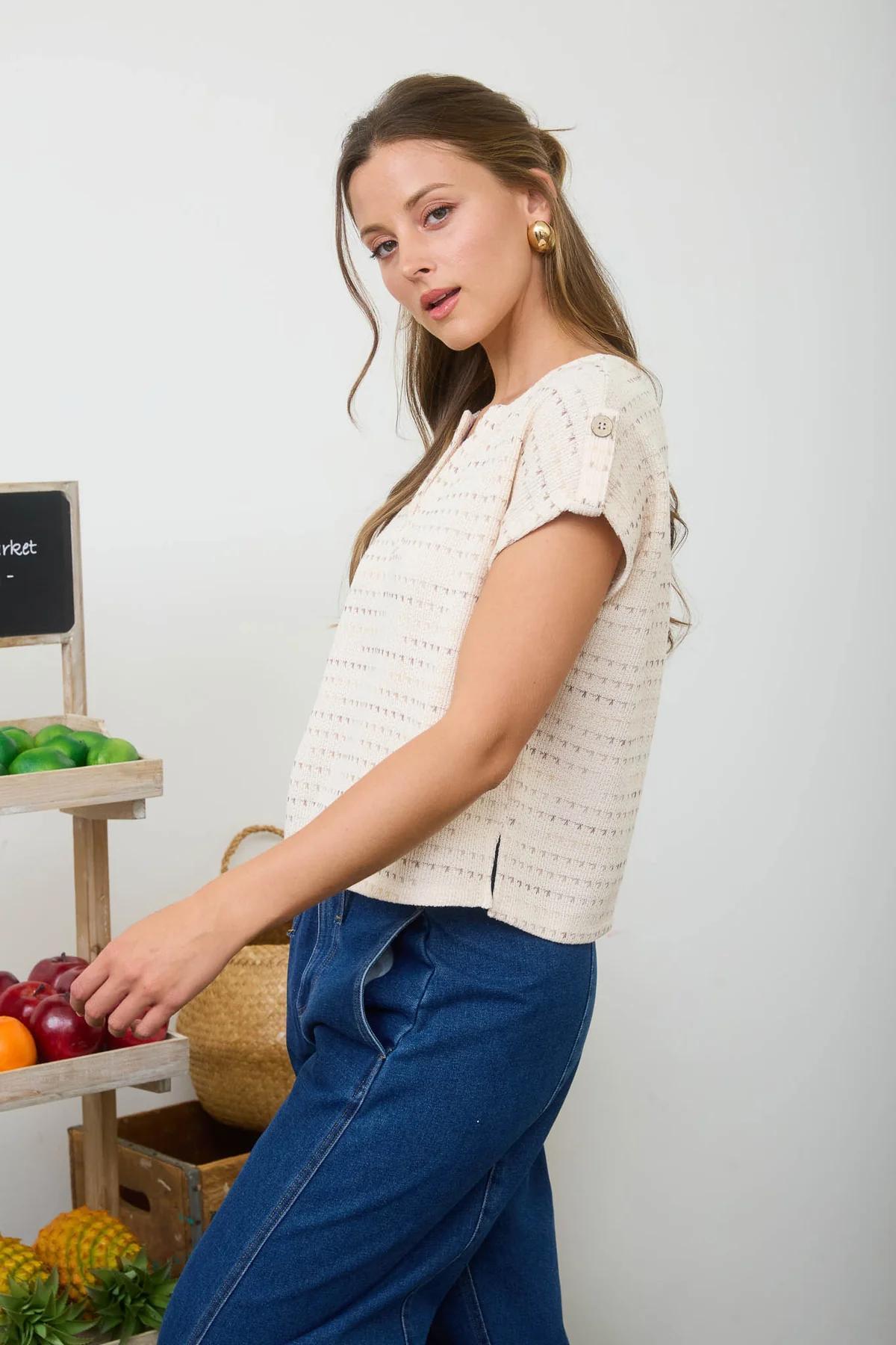 Woman in a white textured top and blue pants standing next to a fruit display.