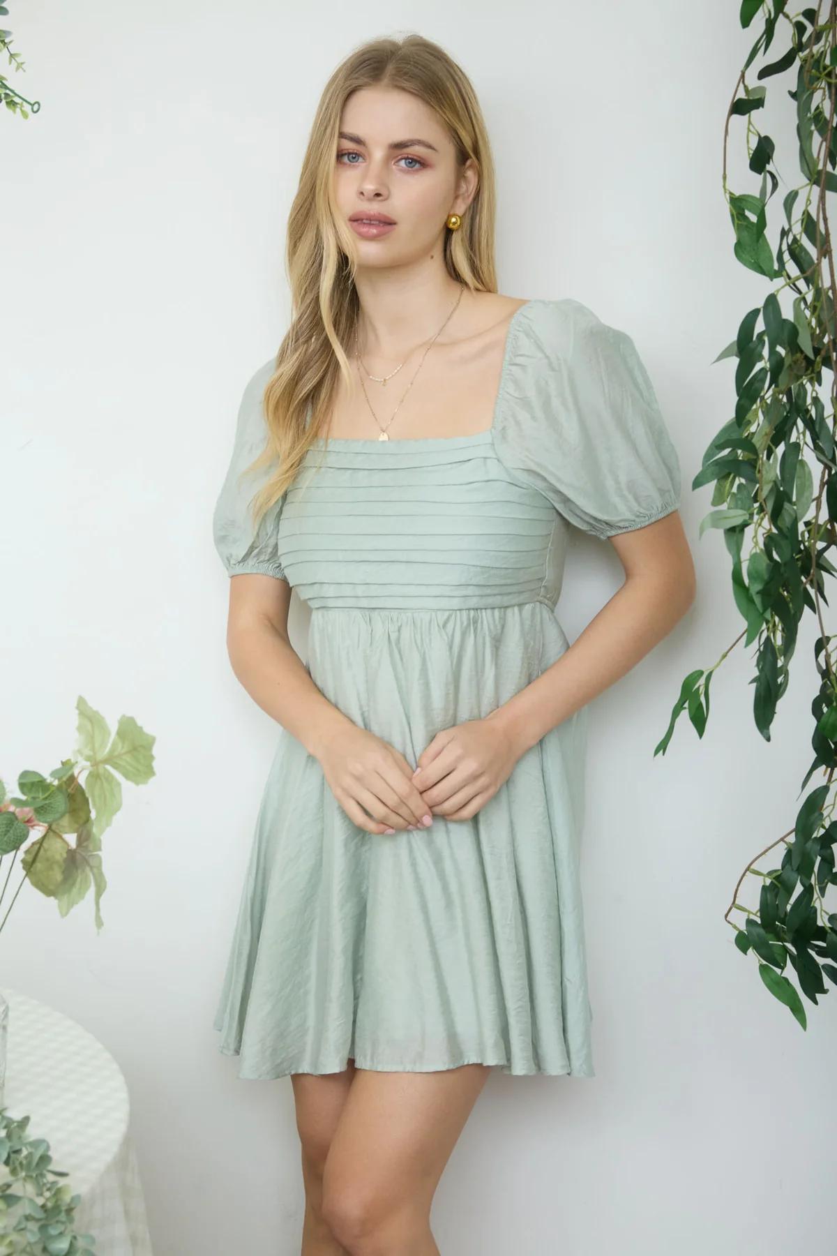 Woman wearing a light green dress standing against a white wall with plants around.