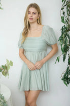 Woman wearing a light green dress standing against a white wall with plants around.